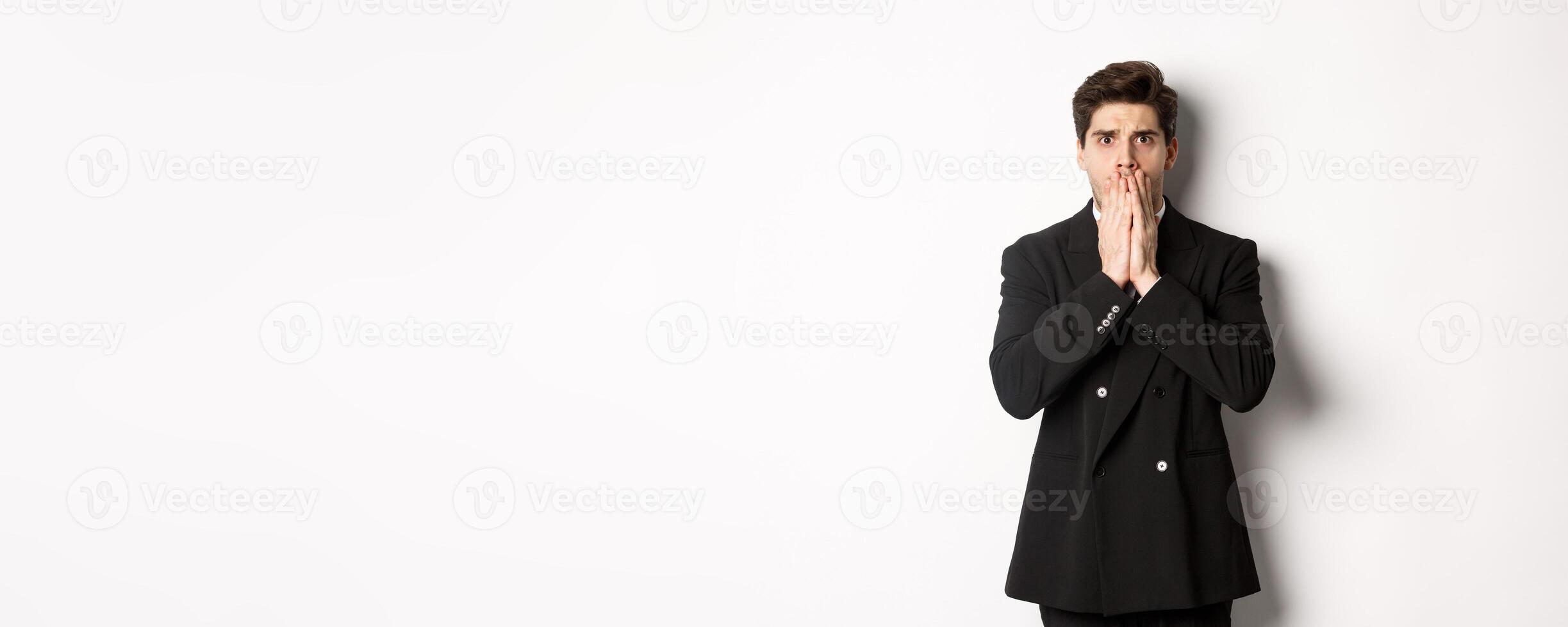 Portrait of shocked handsome businessman in suit, reacting to terrible situation, gasping and covering mouth with hands, standing startled against white background photo