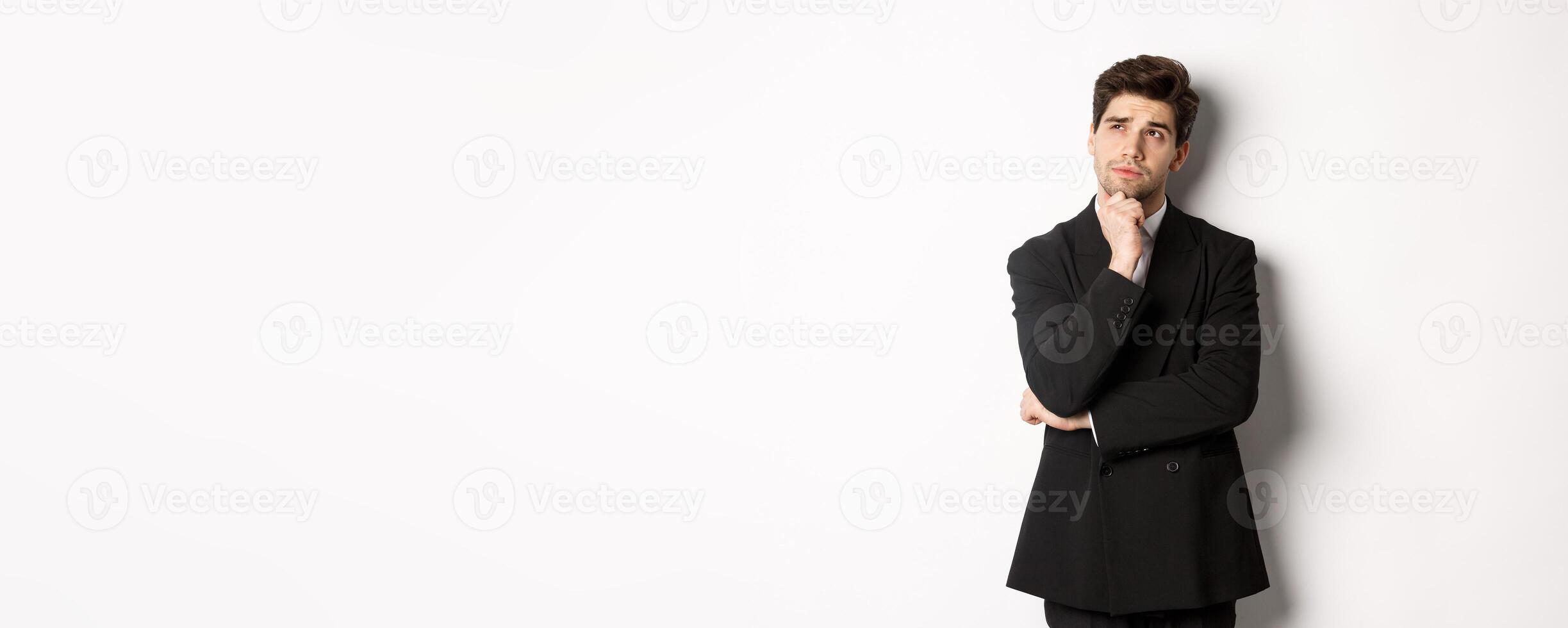 Portrait of handsome and stylish man in suit, thinking and looking at upper left corner, imaging something about christmas holidays, standing over white background photo