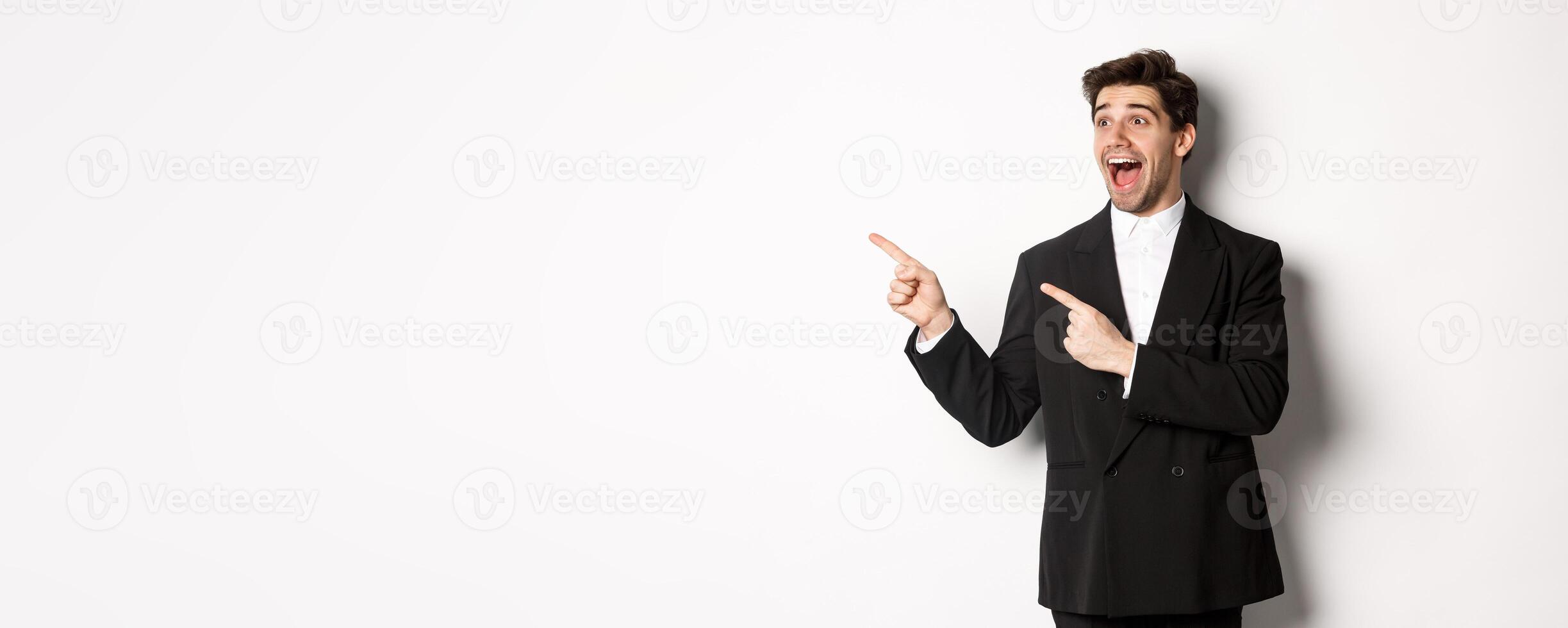 Portrait of amazed and happy handsome man in black suit, pointing and looking left impressed, showing awesome christmas promo, standing over white background photo