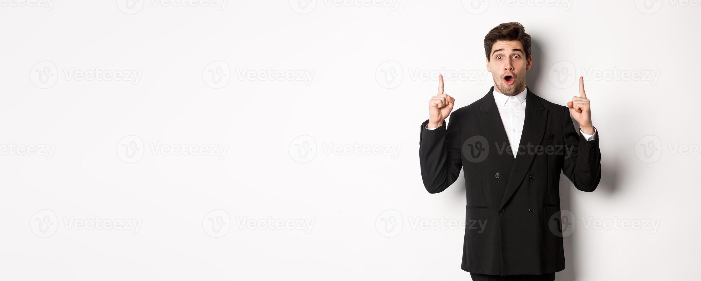 Portrait of surprised handsome businessman in black suit, saying wow and looking amazed, pointing fingers up at copy space, standing over white background photo