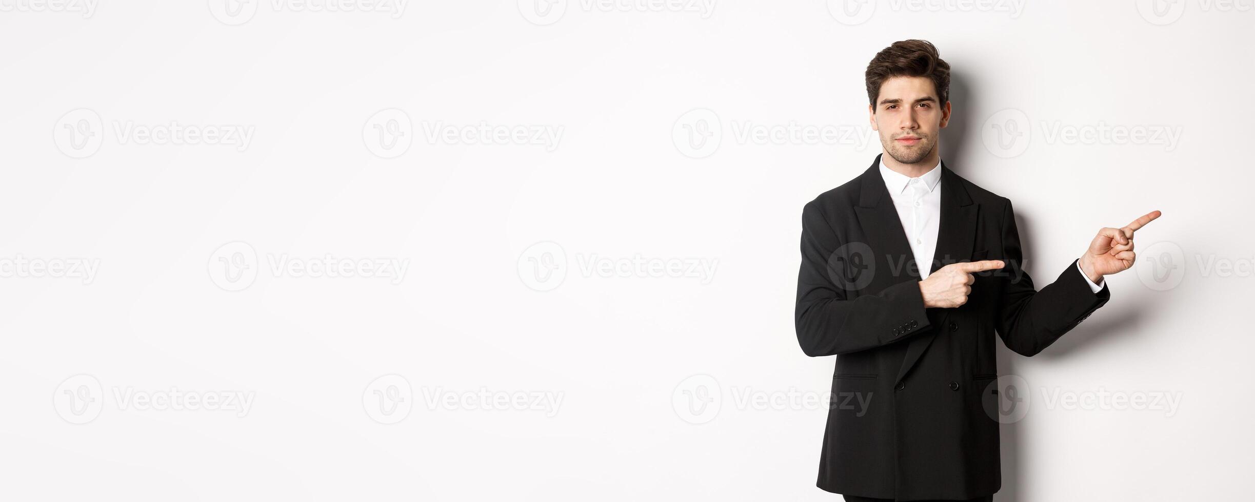 Image of handsome businessman in black suit, pointing fingers right and looking at camera, standing against white background photo