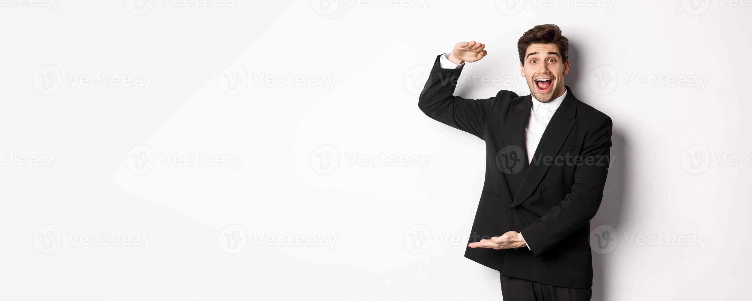 Portrait of excited handsome man in suit, shaping big object on copy space and smiling amazed, holding something, standing over white background photo