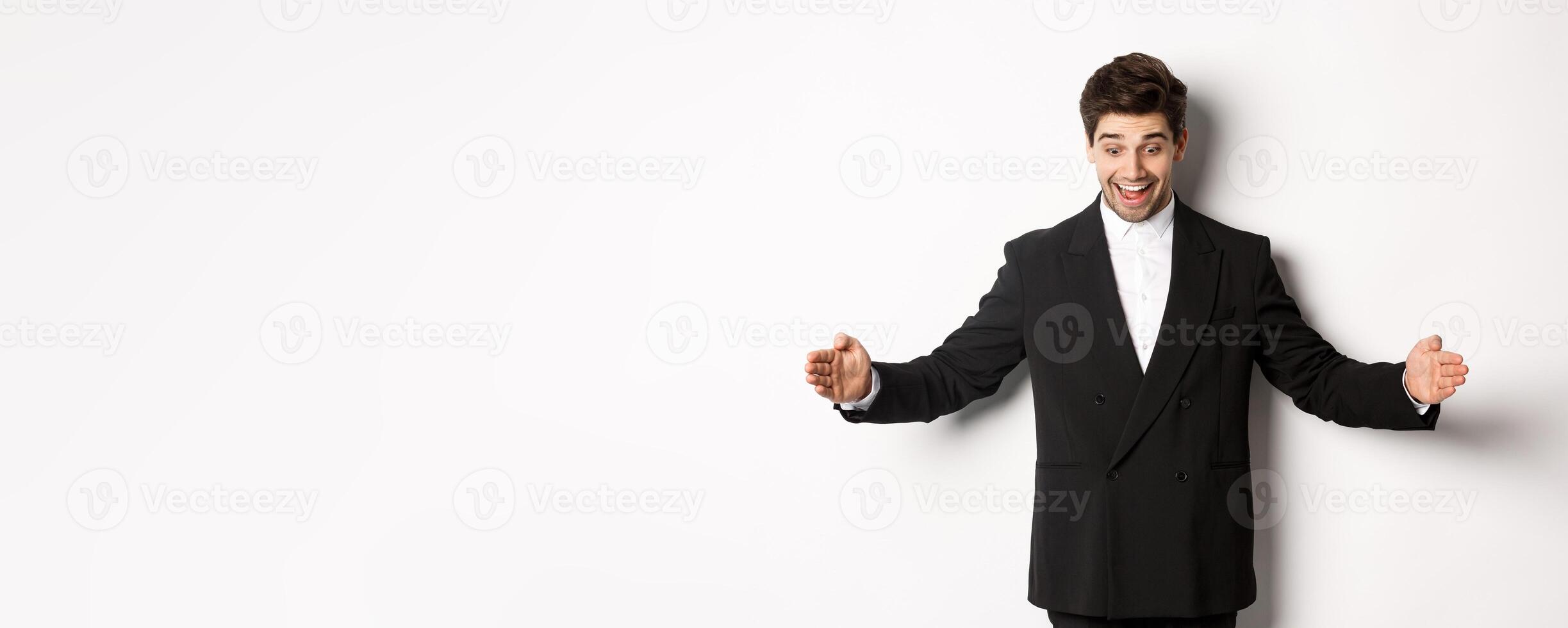 Portrait of excited handsome man in suit, shaping big object on copy space and smiling amazed, holding something, standing over white background photo