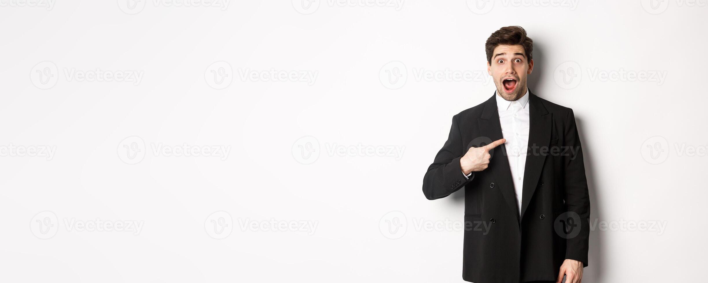 Concept of new year party, celebration and lifestyle. Portrait of surprised handsome guy in black suit, pointing at himself and looking amazed, being chosen, standing over white background photo