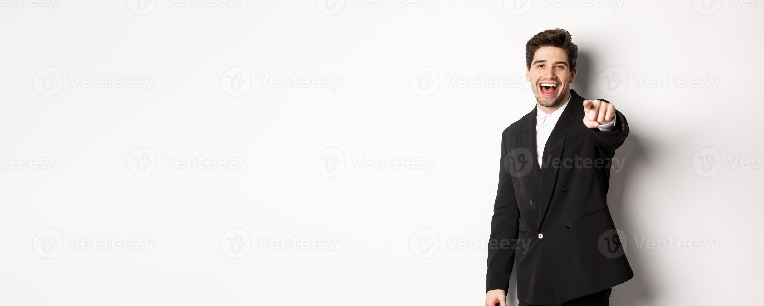 Concept of new year party, celebration and lifestyle. Portrait of handsome stylish man in black suit, smiling and pointing finger at camera, standing over white background photo