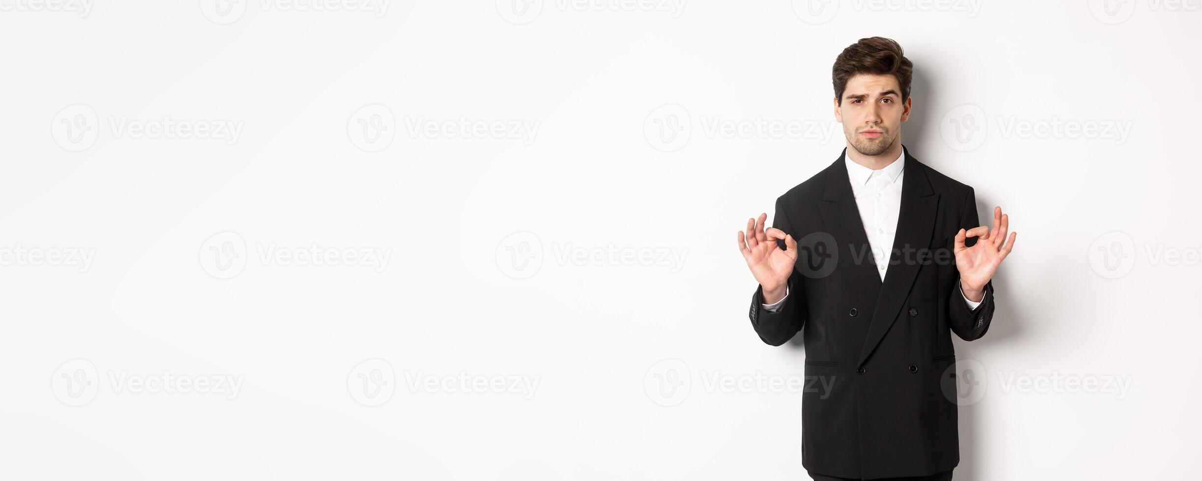 Concept of new year party, celebration and lifestyle. Portrait of confident good-looking man in black suit, showing okay sign and approve something, standing over white background photo