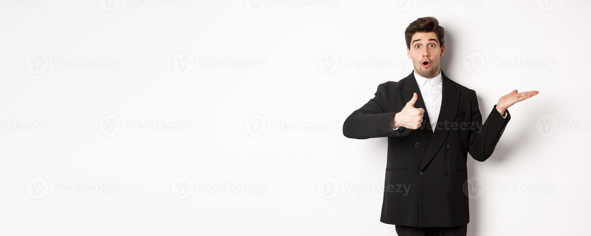 Portrait of hadnsome bearded man in formal suit, showing thumb-up and holding product in hand over white copy space, recommending product, standing over white background photo