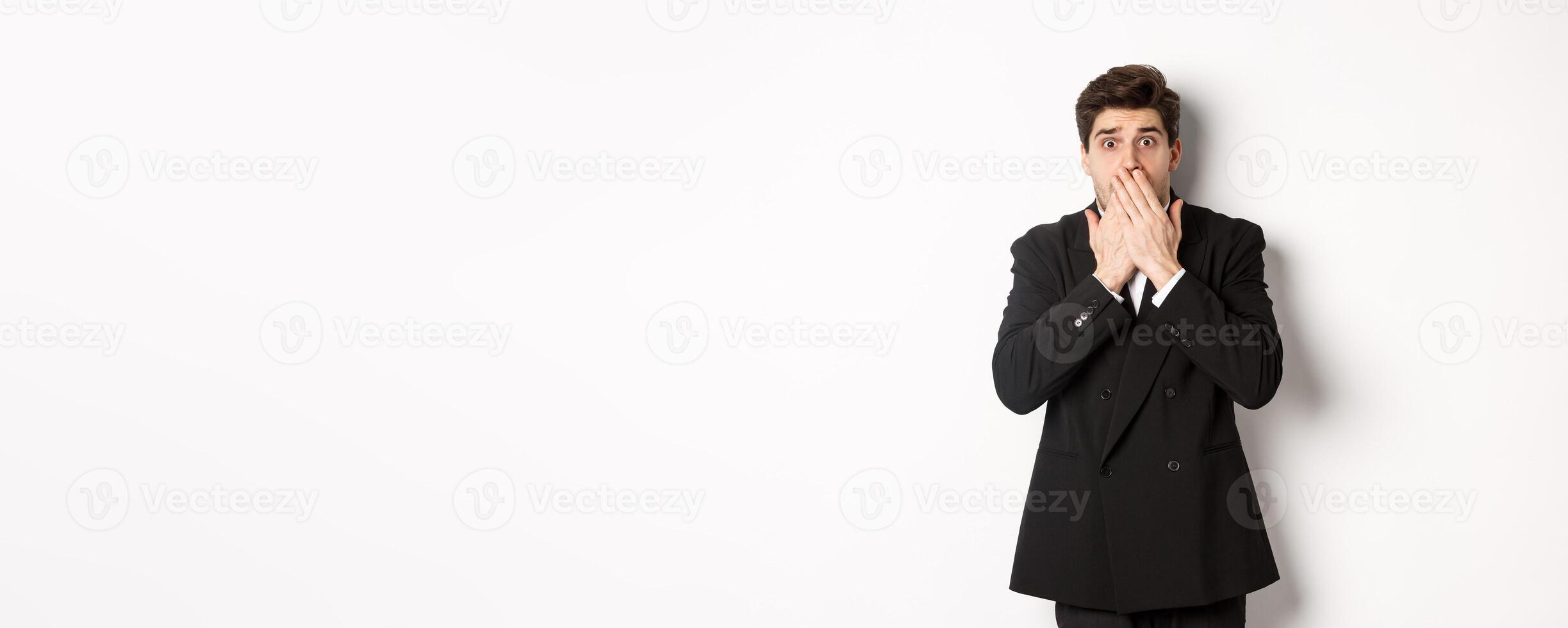 Scared man in formal suit, gasping and looking frightened at camera, standing against white background photo