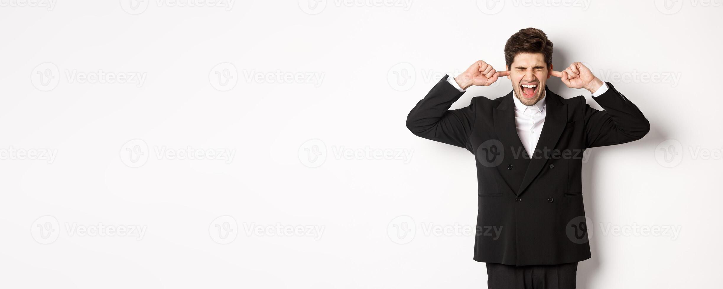 Portrait of annoyed and bothered businessman in black suit, shut ears and yelling, complaining loud noise, standing against white background photo