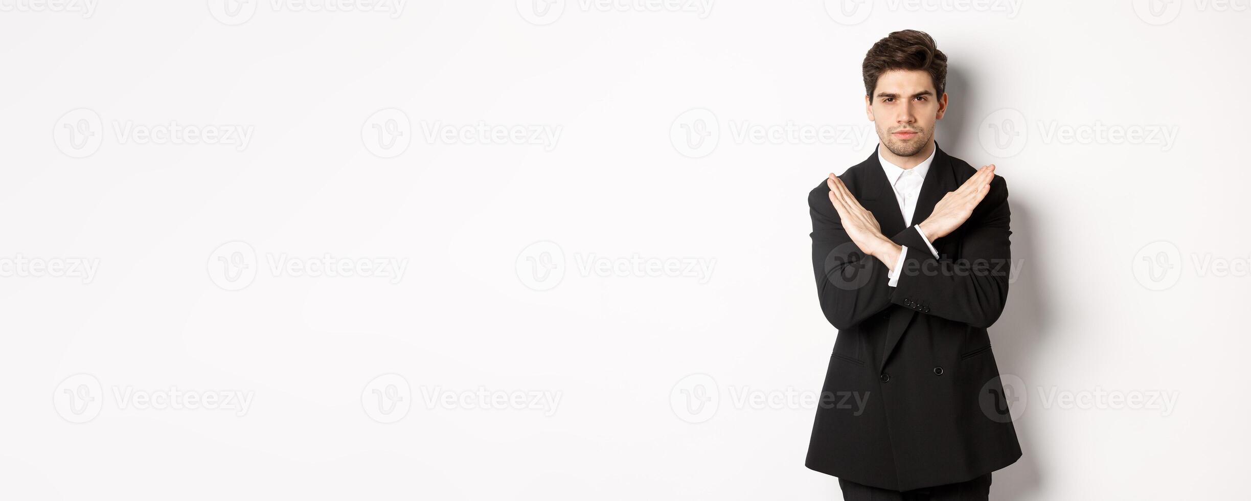 Portrait of serious and concerned man in black suit, showing stop gesture and frowning, making cross to prohibit or decline something, standing over white background photo