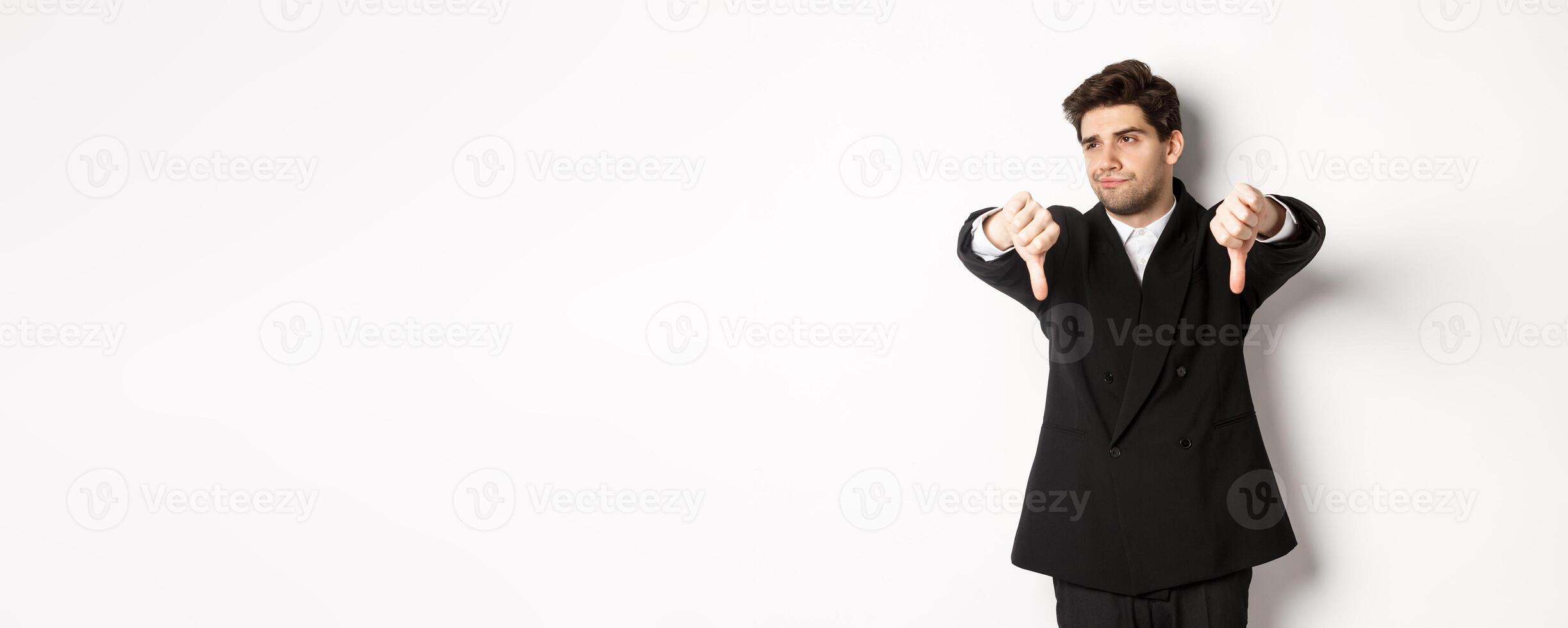 Skeptical and displeased man in black suit, looking away reluctant, showing thumbs-down, dislike something, standing over white background photo