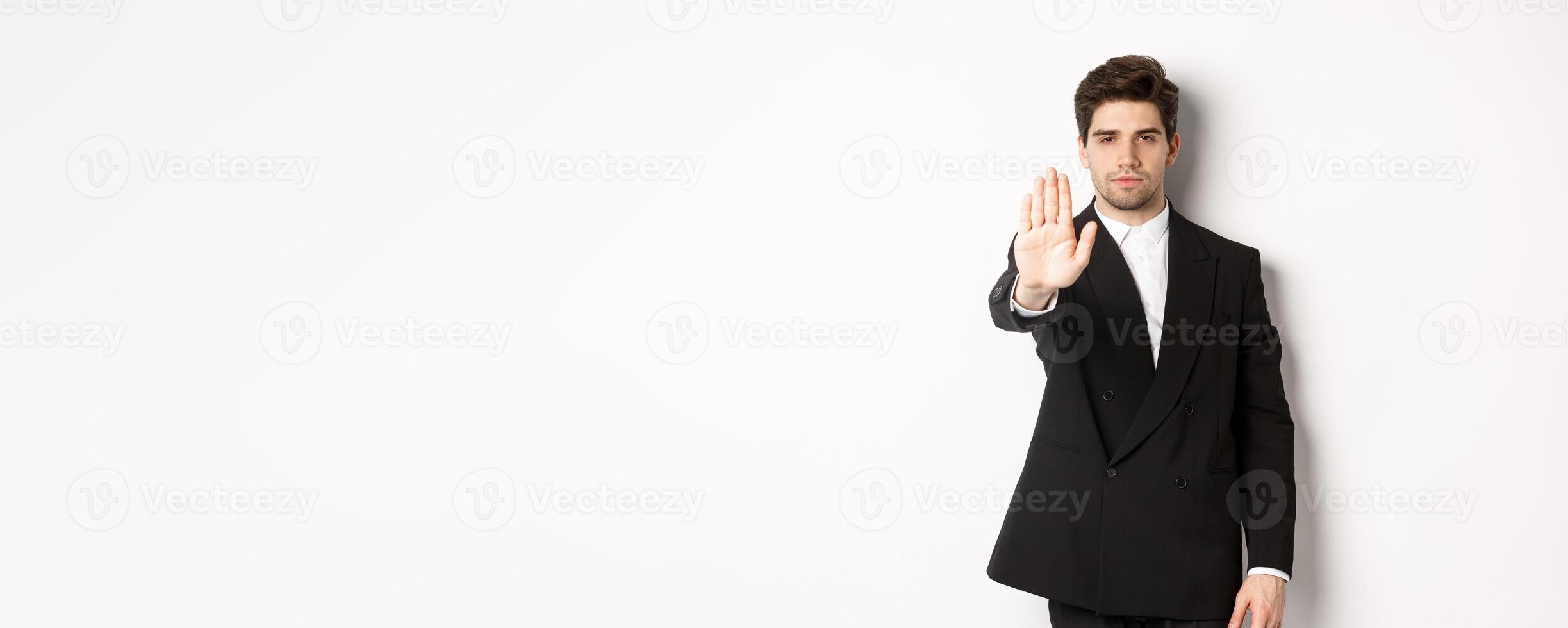 Portrait of serious handsome man in formal suit, extending hand to stop you, prohibit action, forbid and disagree with something, standing against white background photo