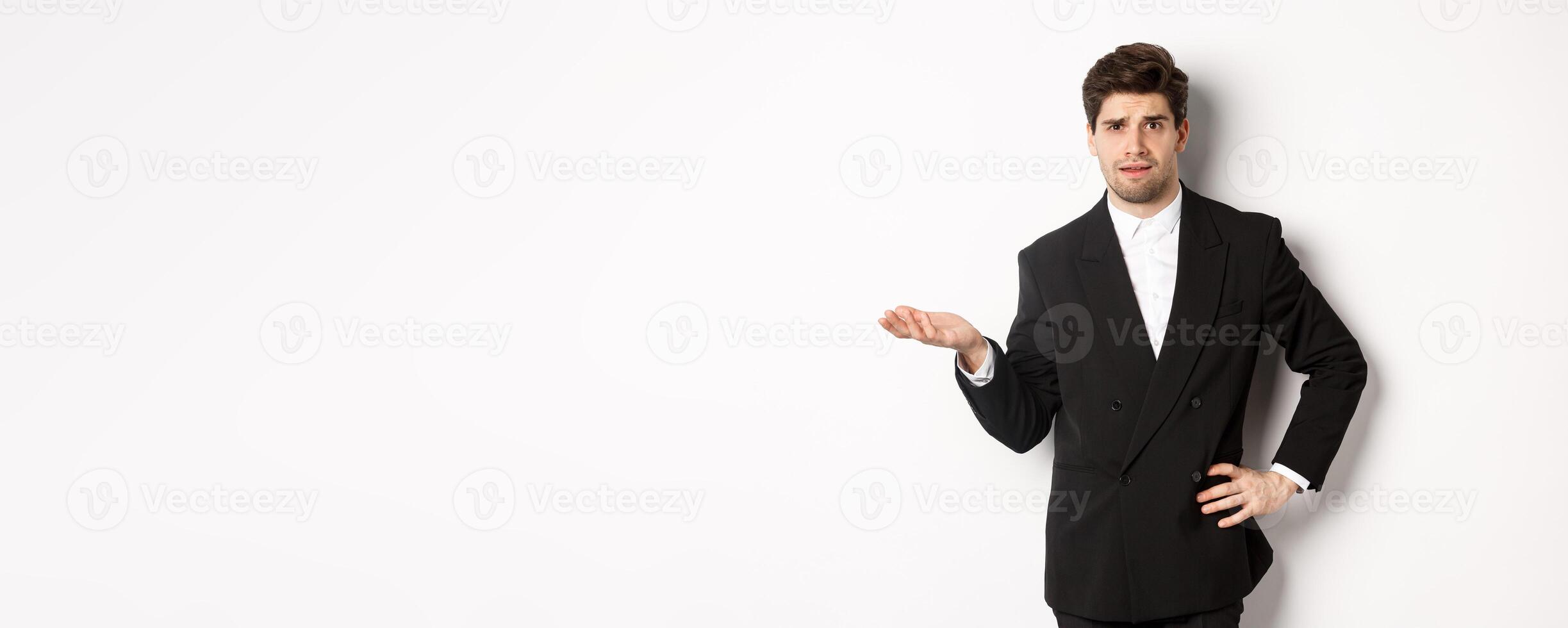 Image of confused handsome man in formal suit, raising hand and shrugging, cant understand something, standing against white background photo