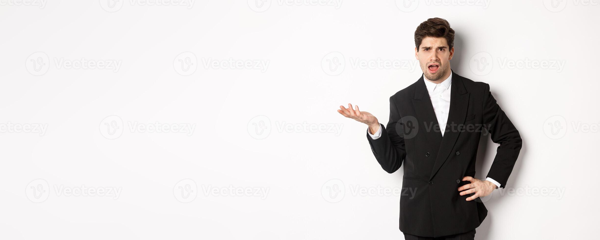 Portrait of arrogant man in black suit, looking confused and disappointed, complaining about something strange, standing over white background photo