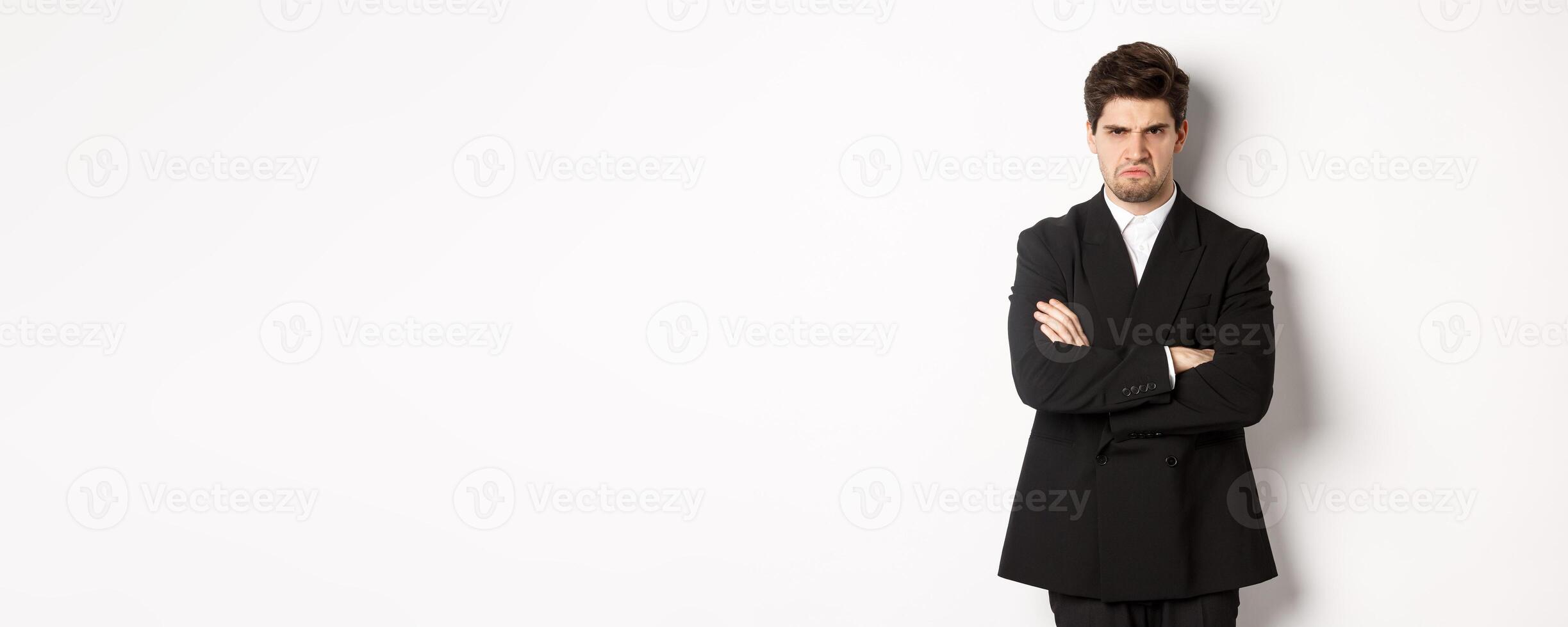 Portrait of angry handsome man in black suit, cross arms on chest and looking offended, frowning and pouting, being mad at someone, standing over white background photo