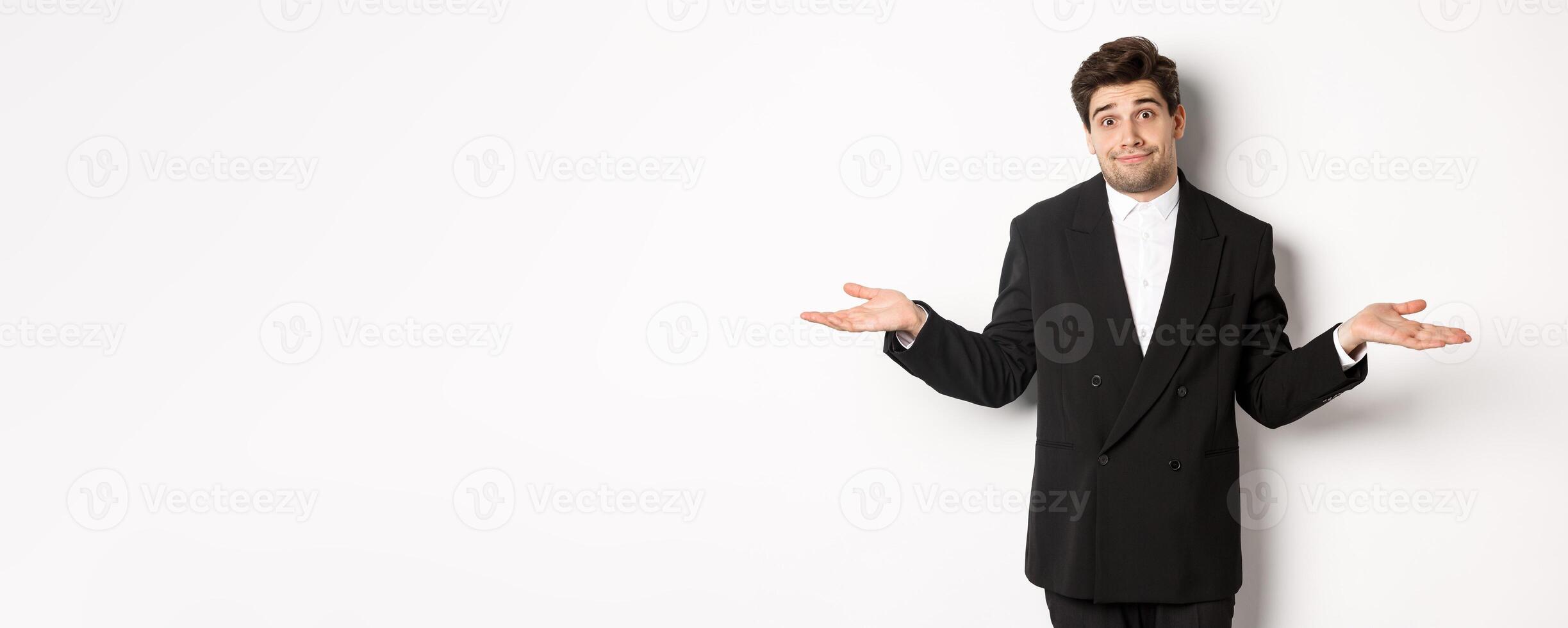 Image of handsome confused man in black suit, shrugging and looking clueless, dont know anything, standing over white background photo