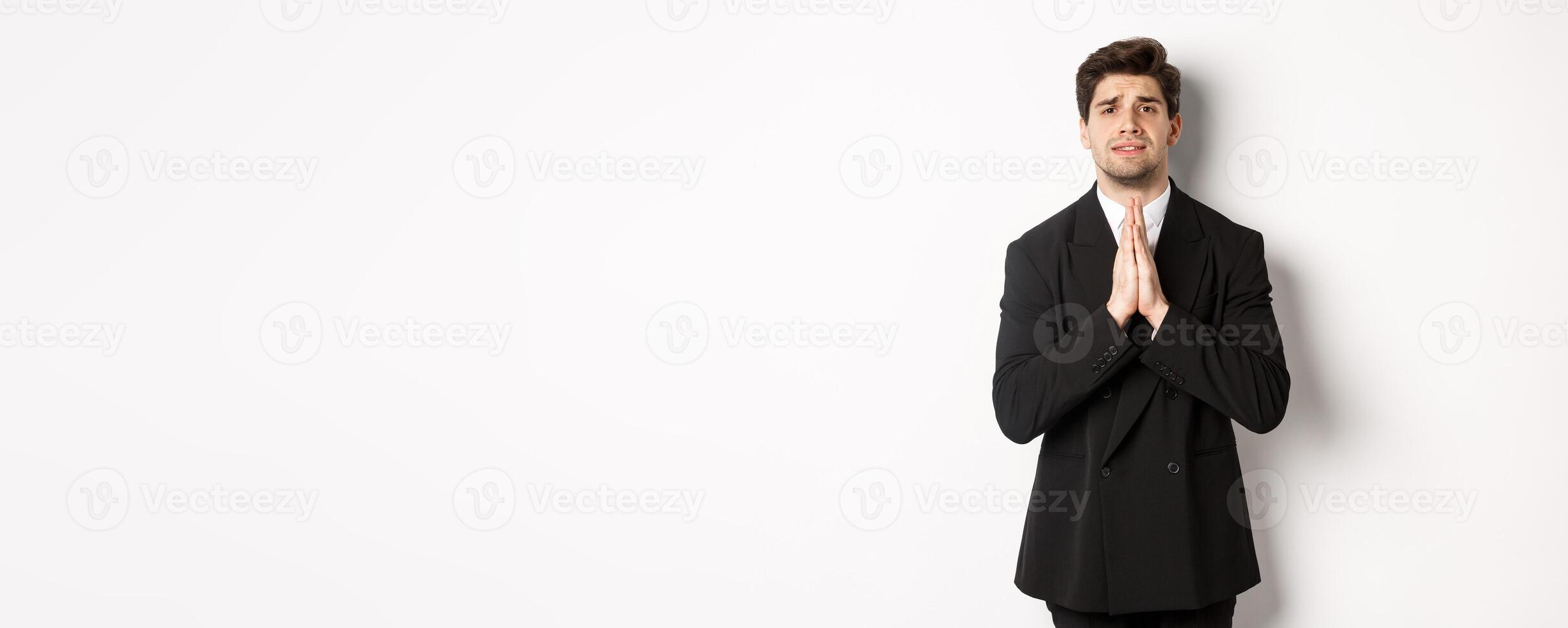 Image of handsome businessman in suit need help, begging you, holding hands in plead gesture and looking desperate, standing over white background photo