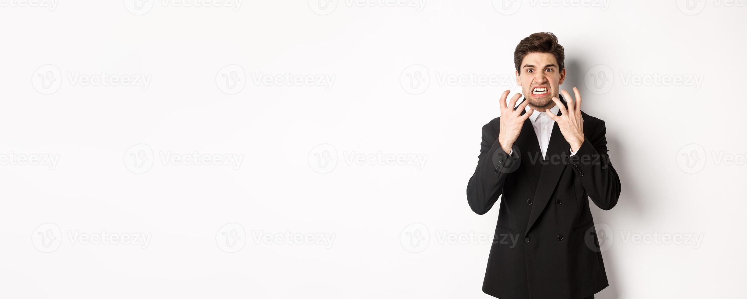 Image of angry businessman in suit, looking with furious expression and clenching fists, express hatred, standing mad over white background photo