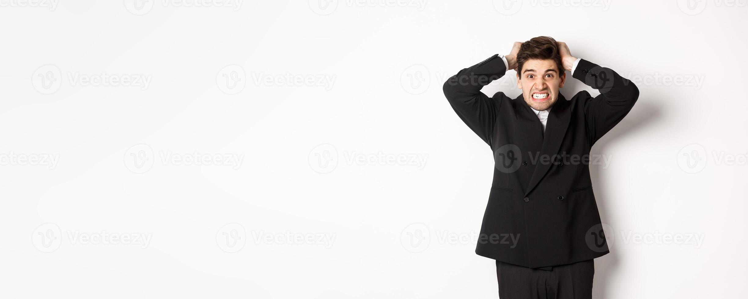 Image of frustrated and angry businessman in black suit, ripping hair on head and grimacing mad, standing tensed against white background photo
