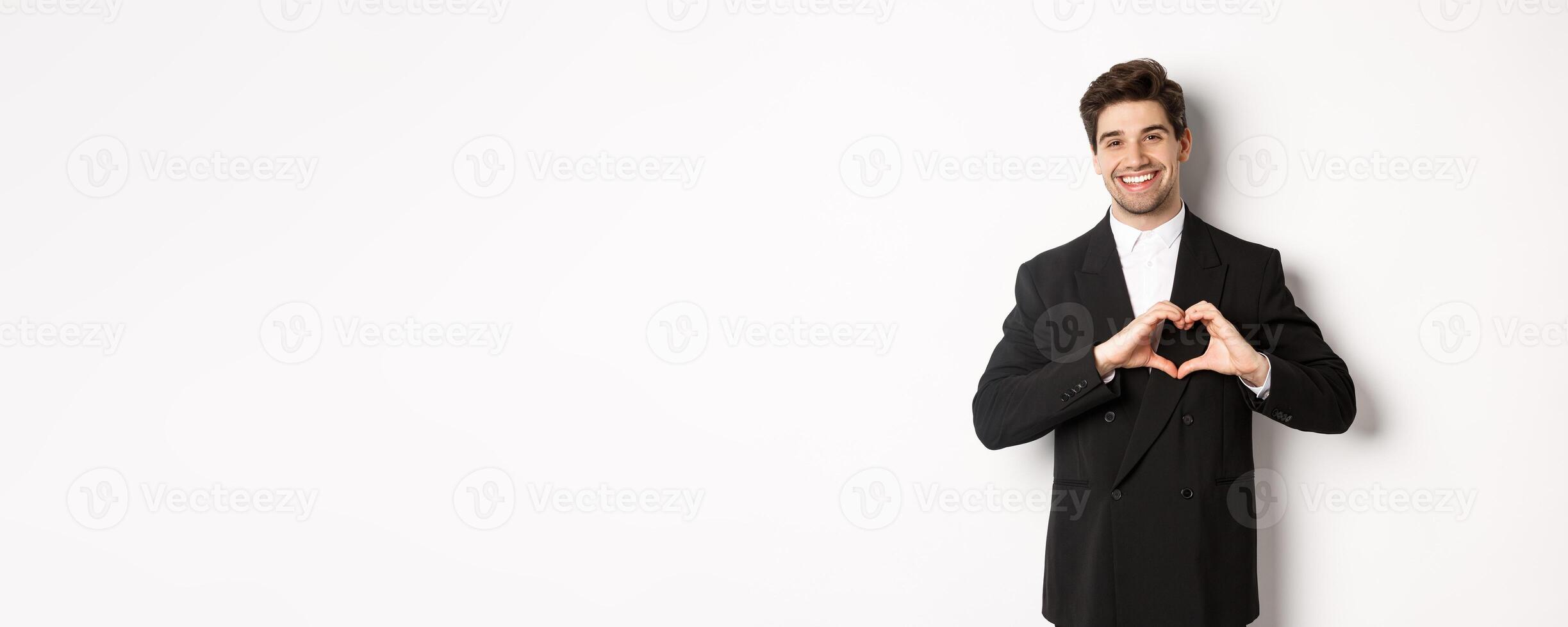 Image of handsome groom in black suit, showing heart sign and smiling, express love and sympathy, standing over white background photo