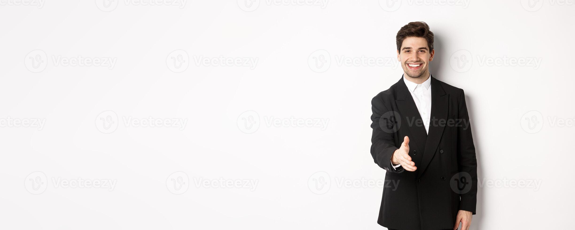 Portrait of handsome businessman in black suit, extending hand for handshake, greet business partners and smiling, welcome to company, standing over white background photo