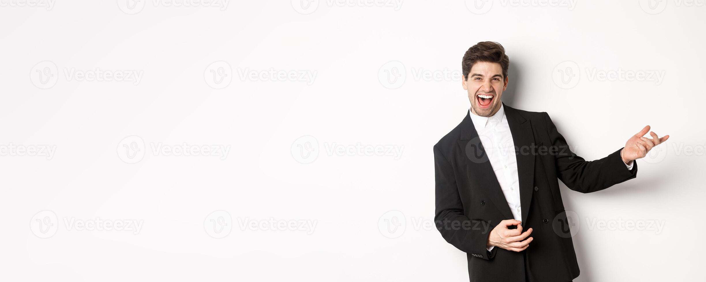 Portrait of happy man dancing at party, playing on invisible guitar and laughing, standing in black suit against white background photo