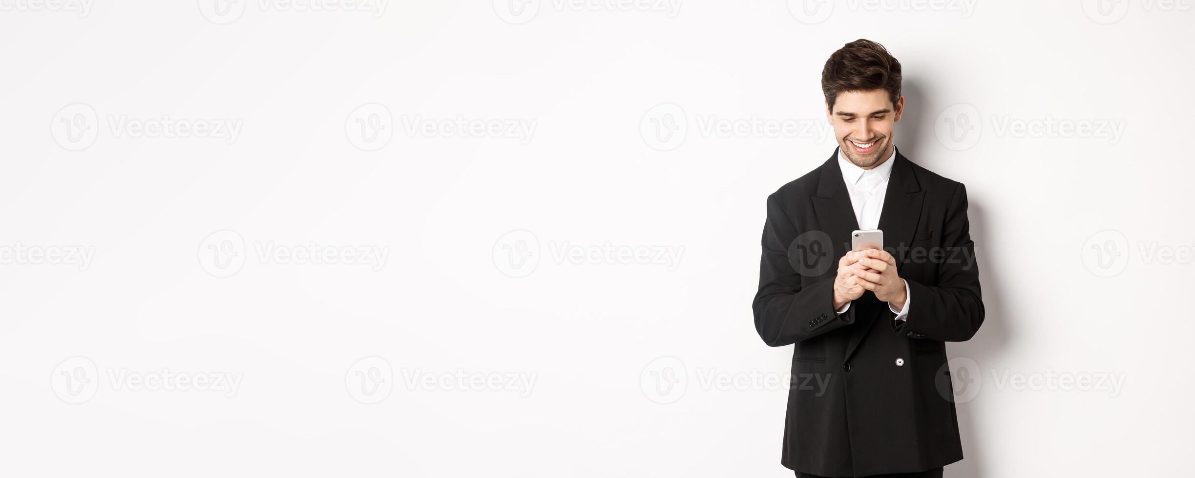 Portrait of handsome stylish businessman in black suit, writing a message, smiling and looking at smartphone, standing over white background photo