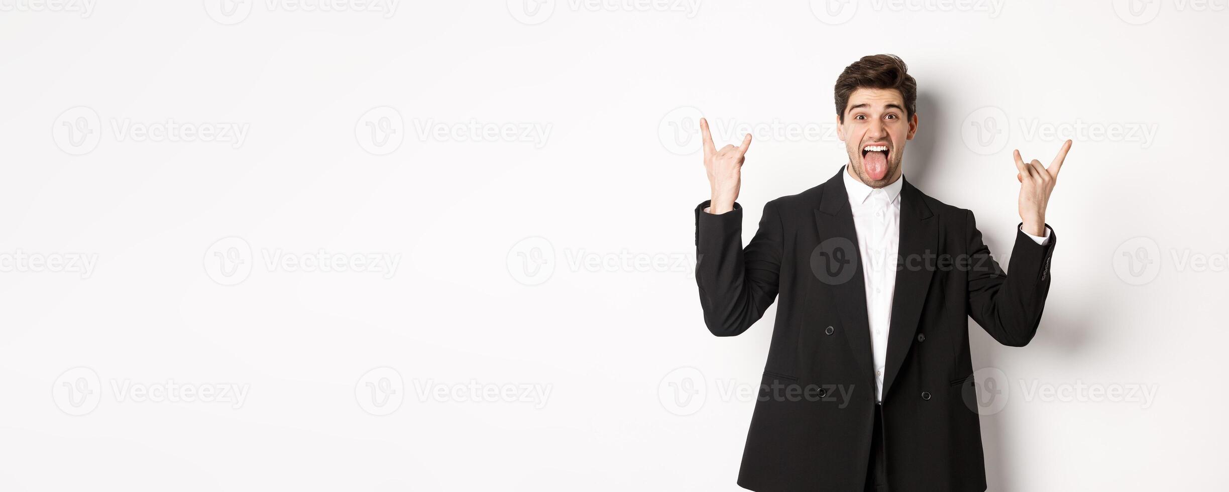 Portrait of happy attractive guy having fun at party, wearing black suit, showing rock-n-roll sign and tongue, standing excited against white background photo