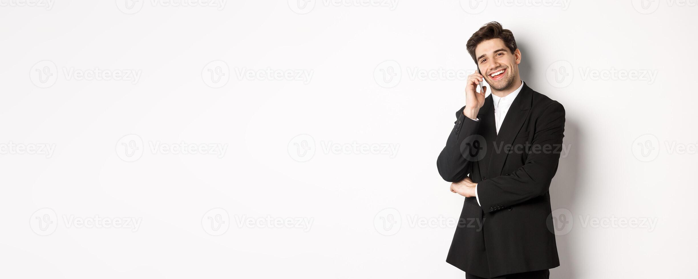 Image of handsome, successful businessman talking on phone, smiling pleased, standing in suit against white background photo