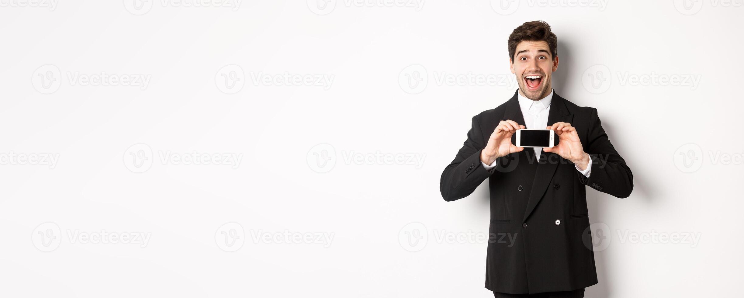 Image of cheerful, handsome man in black suit, showing smarthone screen and looking amazed, standing against white background photo