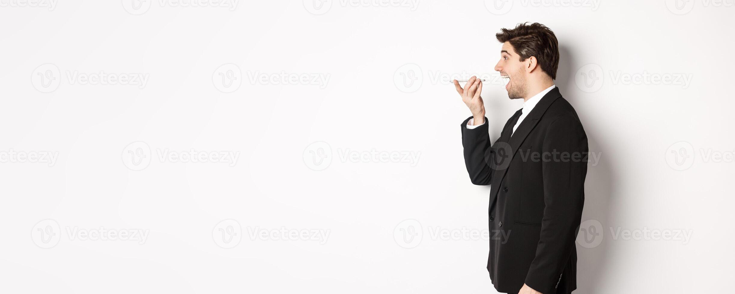 Profile shot of handsome businessman in black suit talking on speakerphone, smiling and looking happy, recording voice message, standing over white background photo
