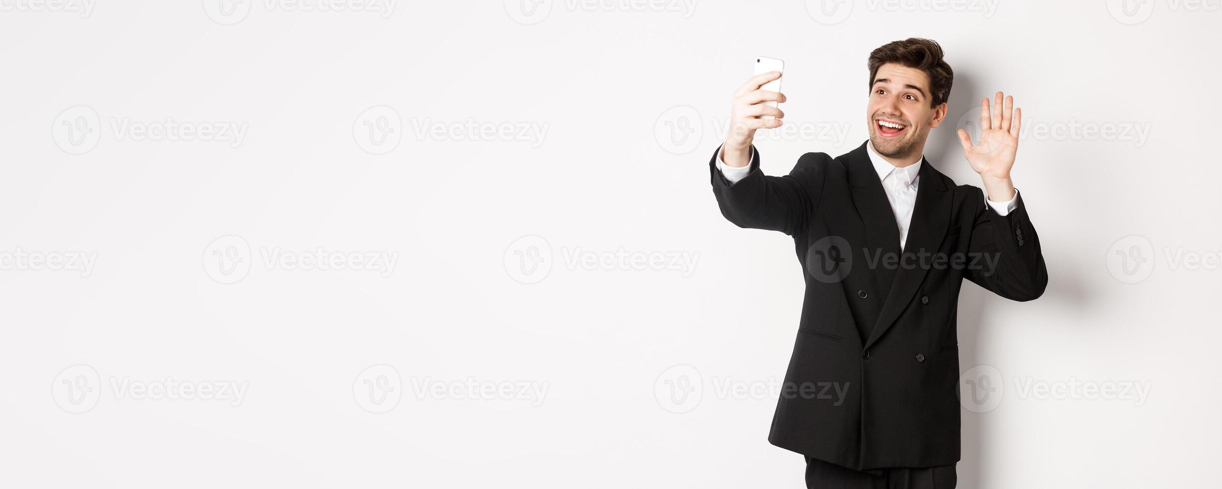 Image of handsome man in suit, having call and waving hand at smartphone camera, recording , greeting someone, standing against white background photo
