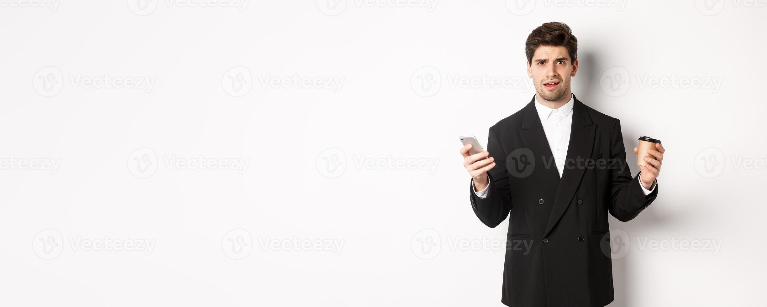 Image of confused businessman standing in a suit, holding cup of coffee and mobile phone, cant understand something, standing against white background photo