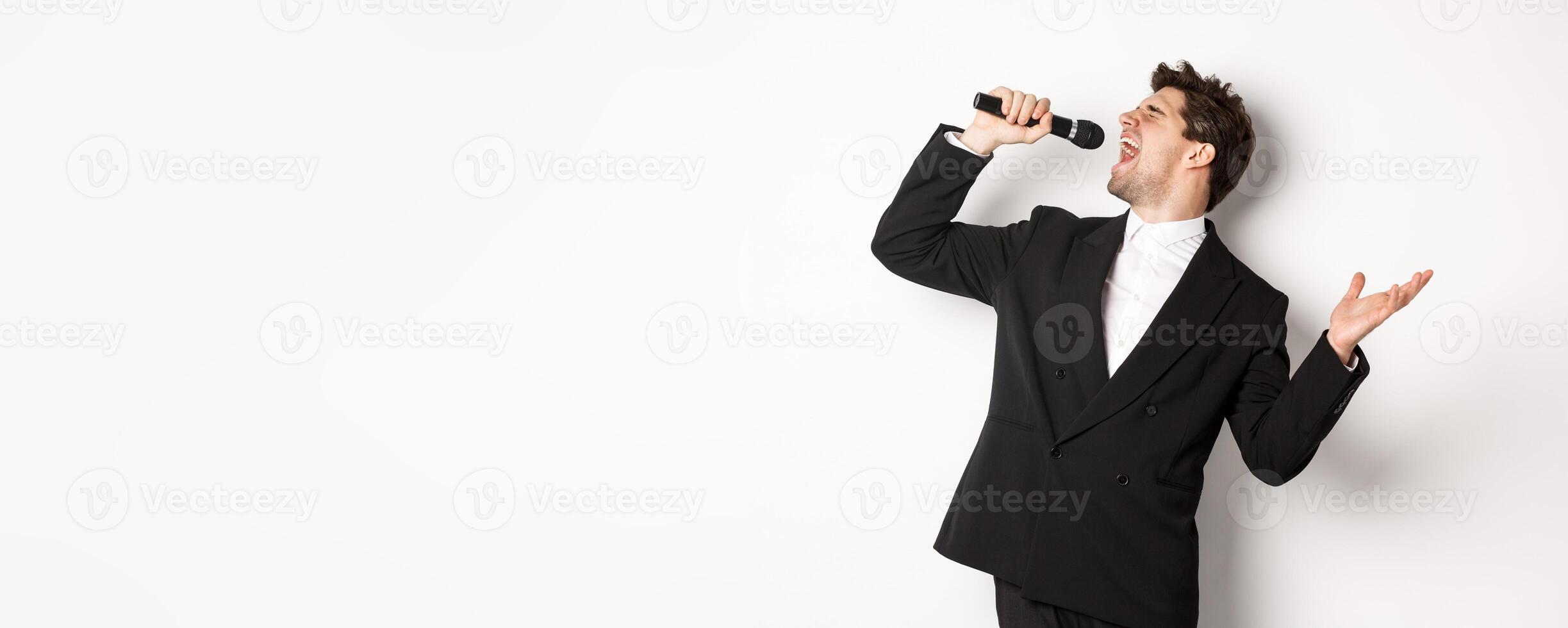 Portrait of handsome man singing a song with passion, standing in black suit, holding microphone and performning, posing over white background photo