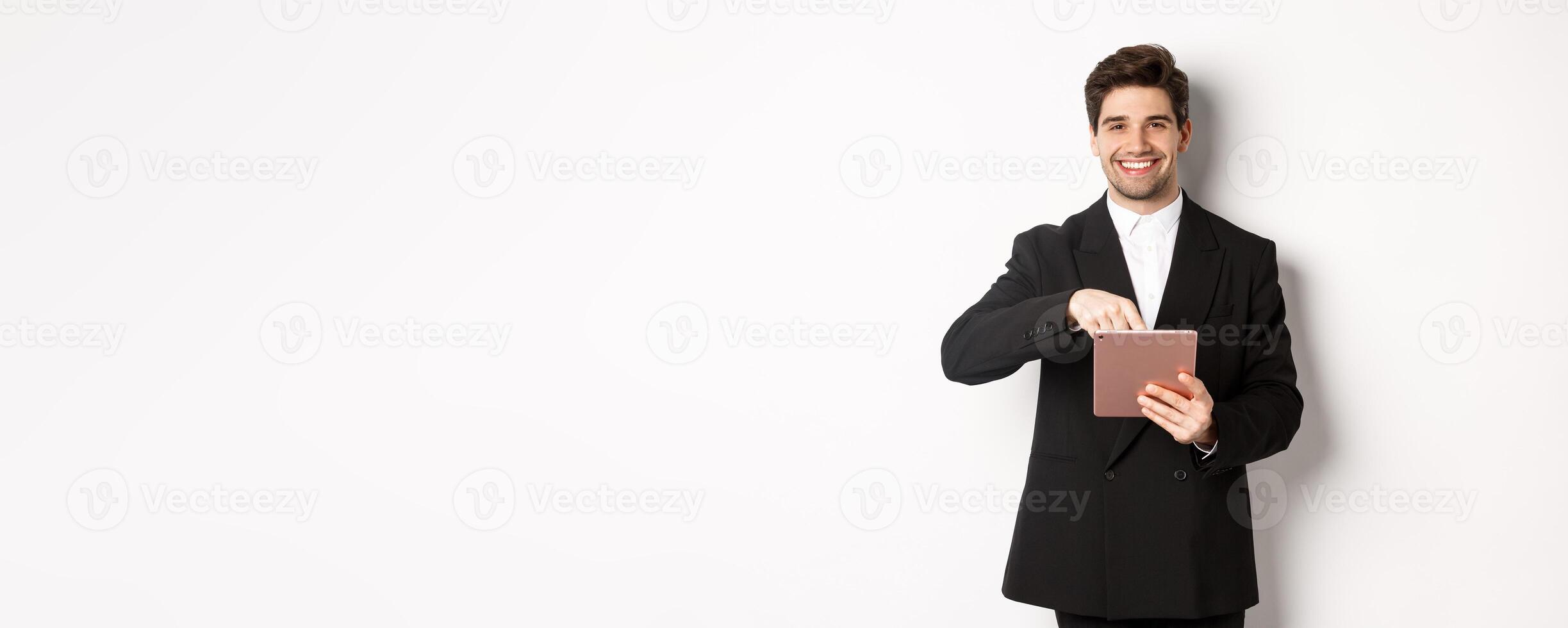 Portrait of handsome, stylish male entrepreneur in black suit pointing at digital tablet, showing something online, standing against white background photo
