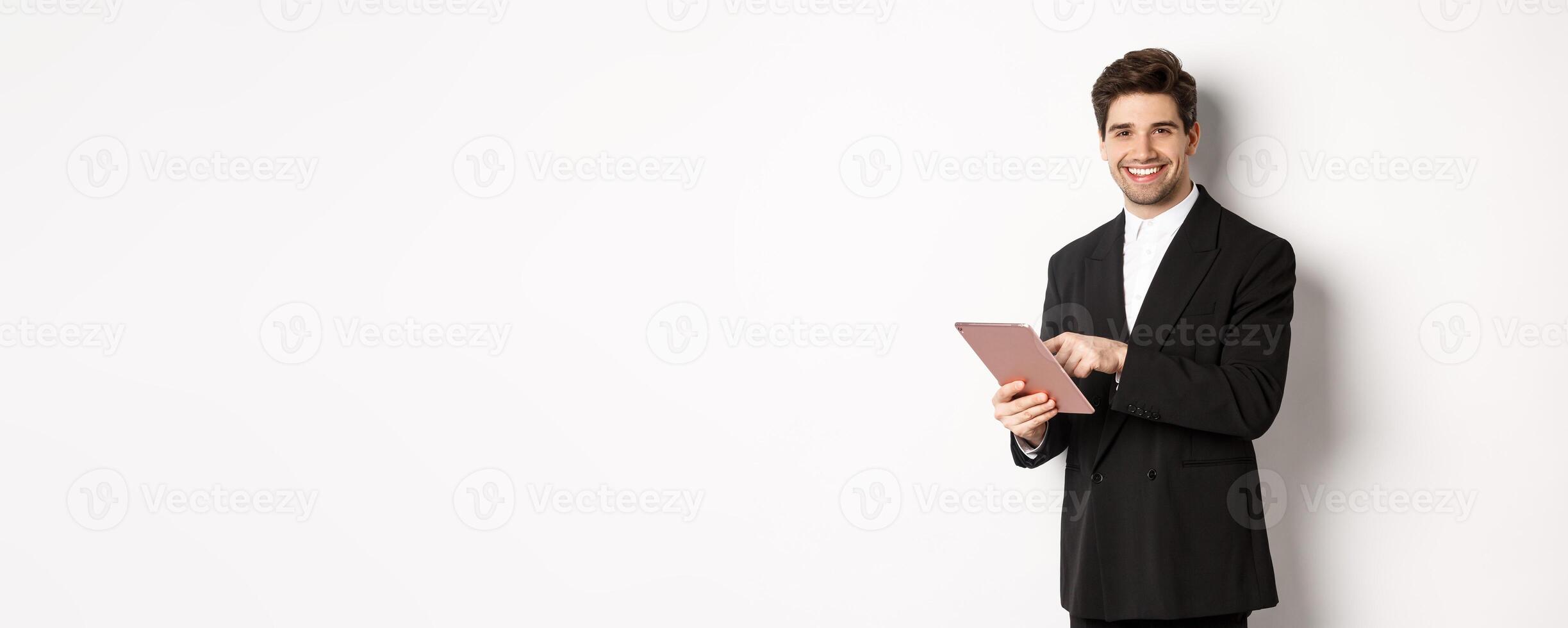 Image of handsome businessman in black suit, using digital tablet and smiling, standing against white background photo