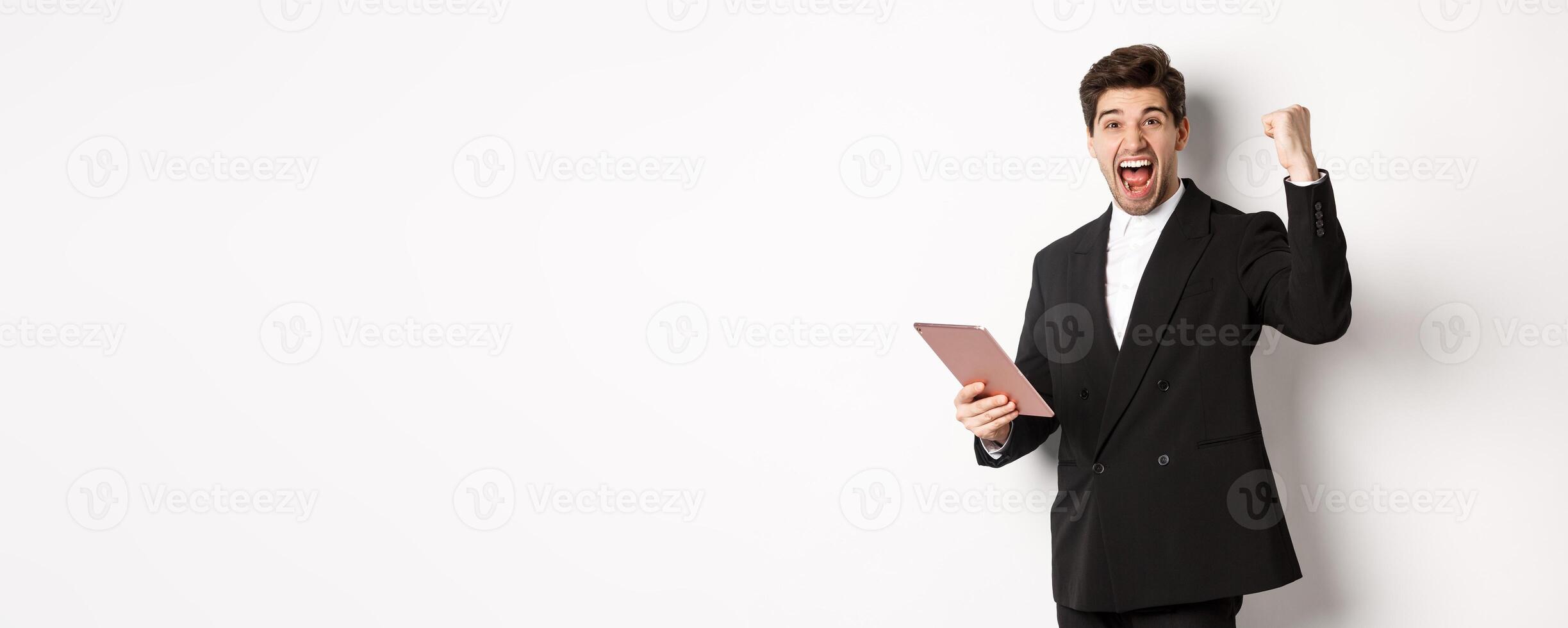 Portrait of handsome successful businessman in black suit, achieve goal, holding digital tablet and saying yes, standing against white background and rejoicing photo