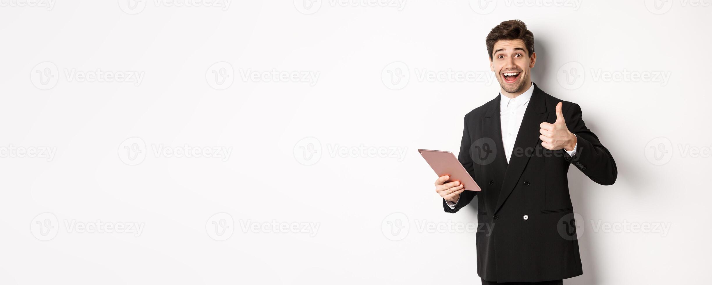 Portrait of handsome businessman in suit, showing thumb-up and making a compliment, praising something on digital tablet, standing over white background photo