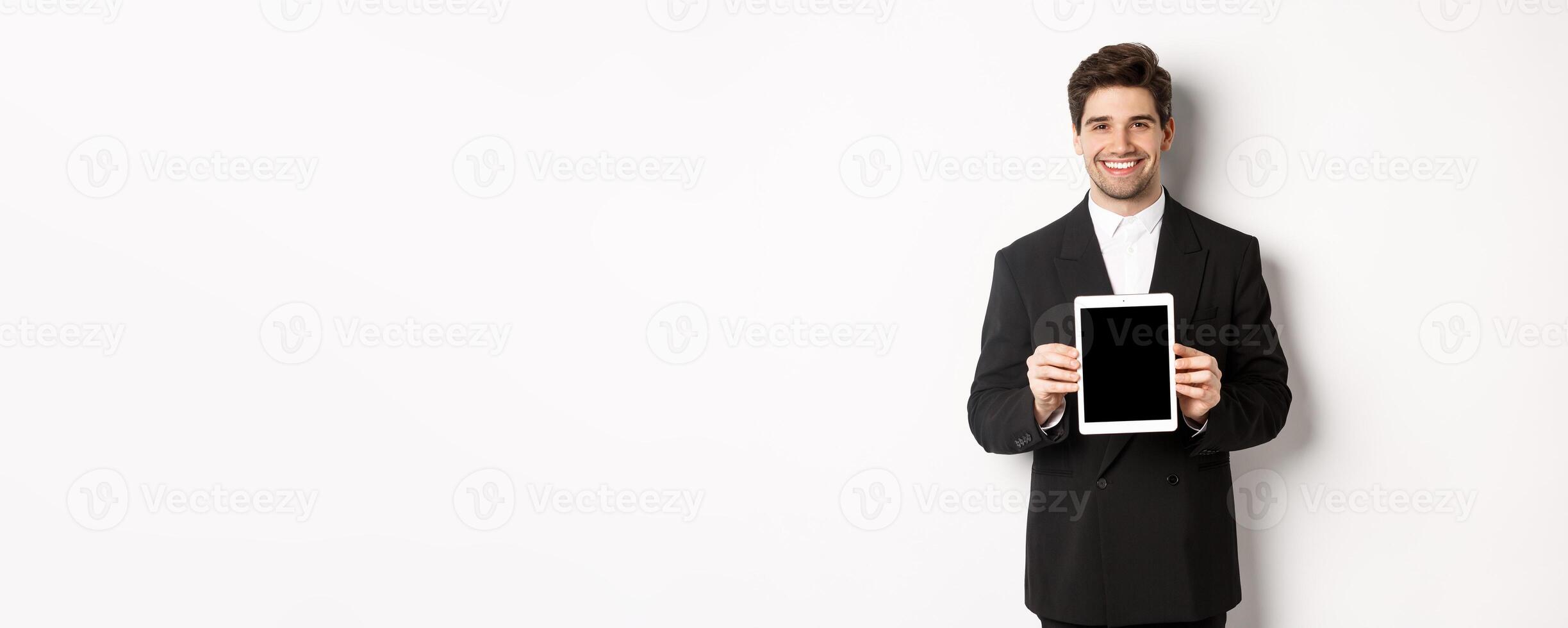 Portrait of handsome businessman in trendy suit, showing digital tablet screen and smiling, standing against white background photo