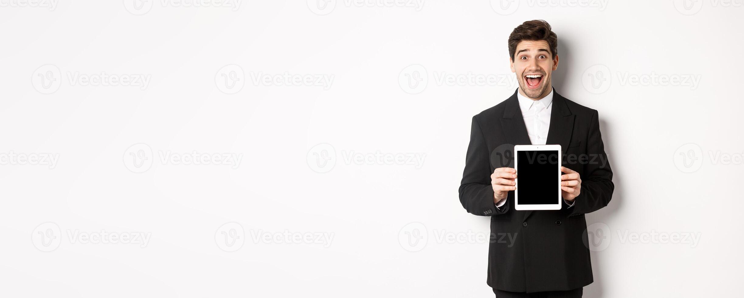 Image of attractive male entrepreneur in trendy suit, showing digital tablet screen and smiling amazed, standing over white background photo