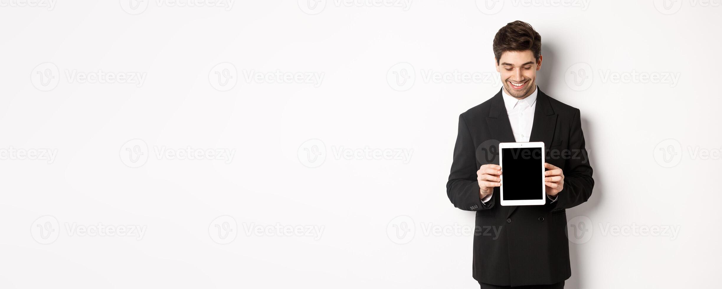 Image of good-looking male entrepreneur in black suit, looking down at digital tablet screen and showing advertisement, standing against white background photo