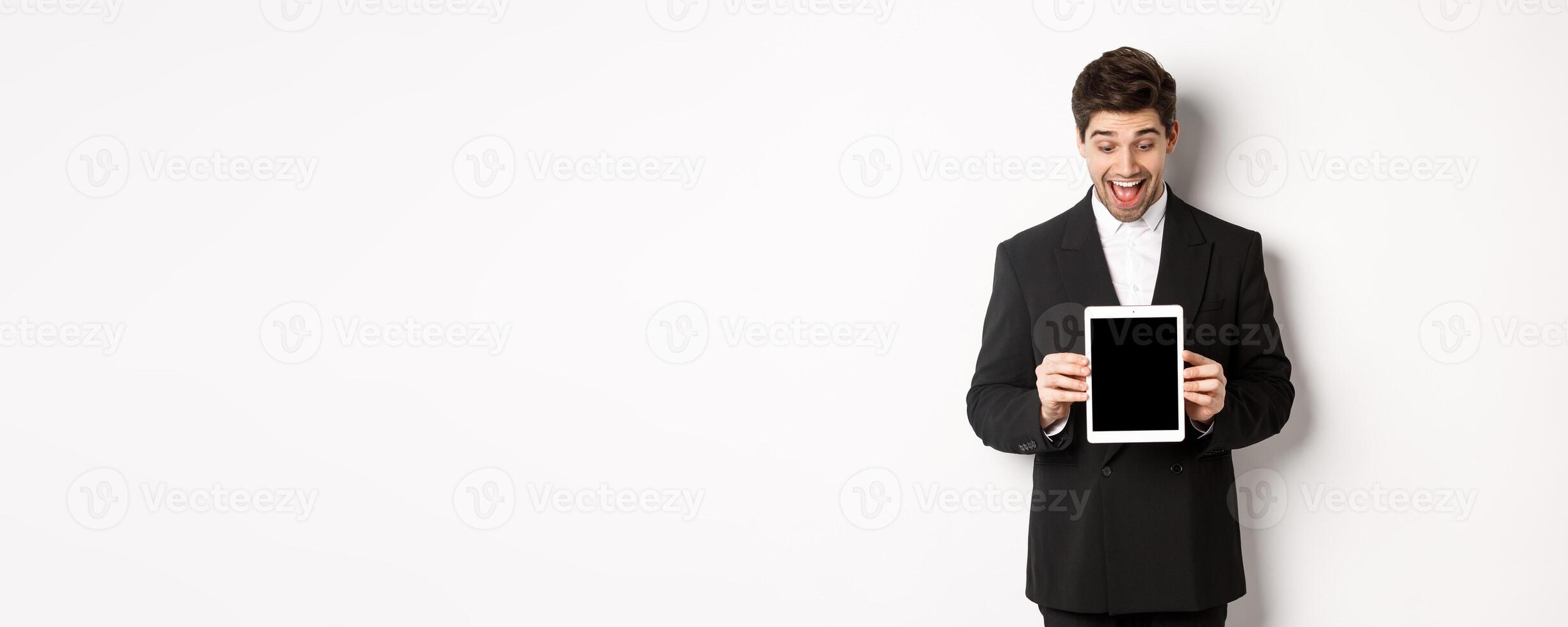 Portrait of amazed handsome man in black suit, showing digital tablet screen with excitement, standing over white background photo