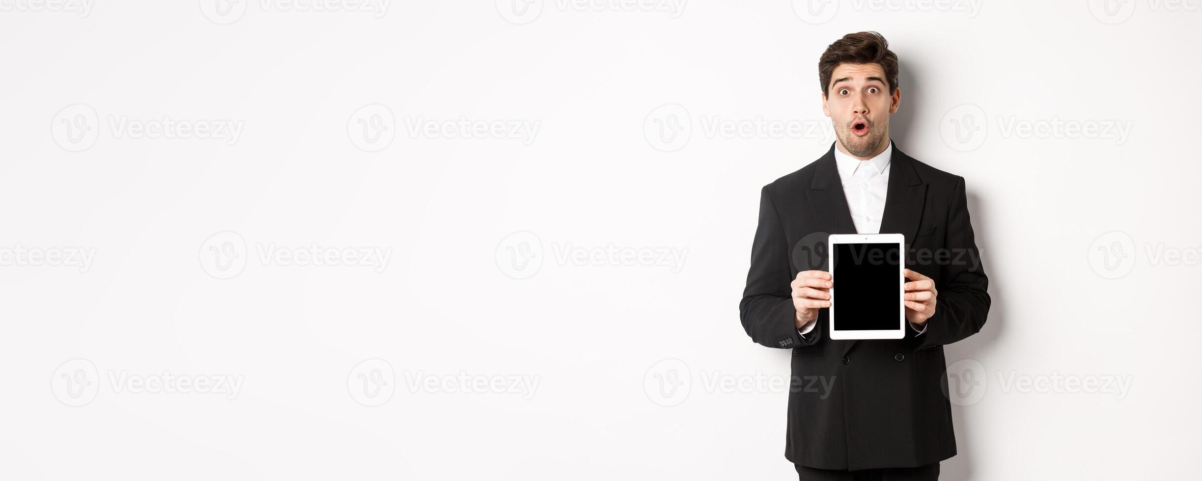 Image of surprised handsome man in black suit, showing digital tablet screen and looking amazed, standing against white background photo