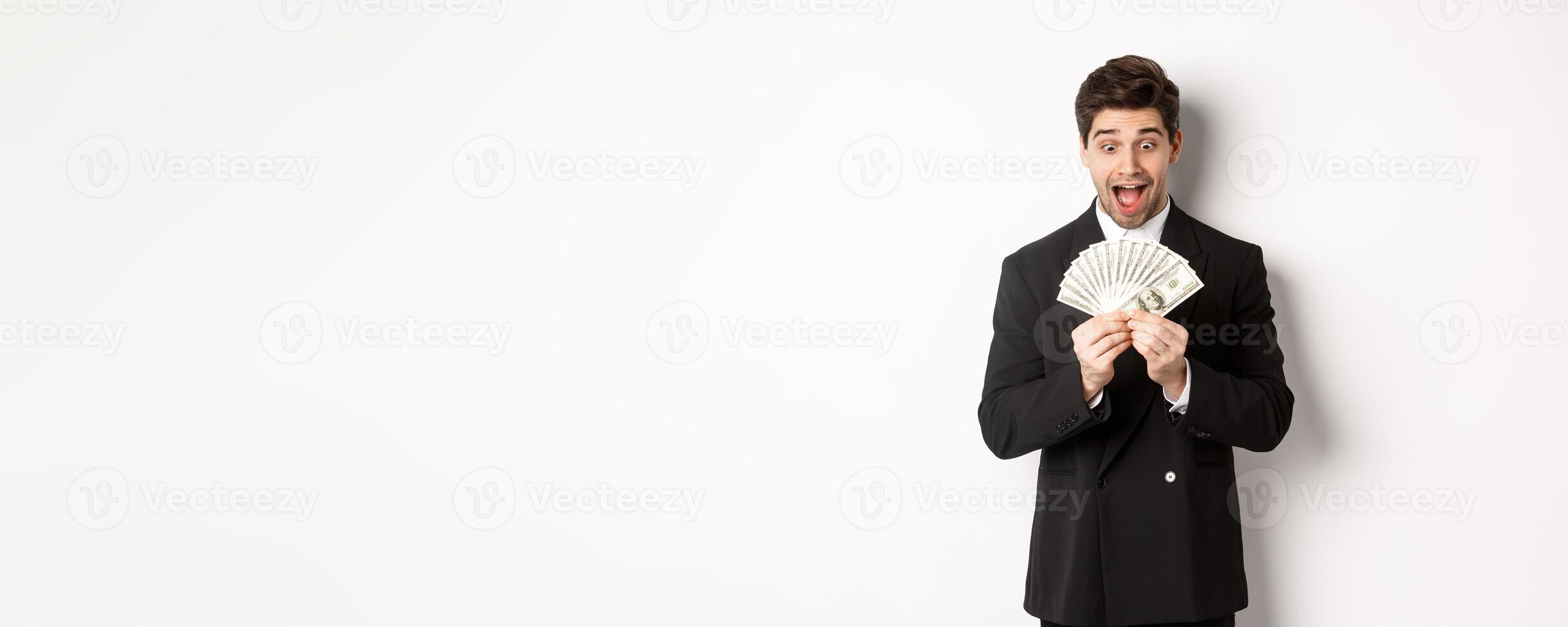 Image of handsome bearded guy in black suit, looking at money with excitement, standing over white background photo