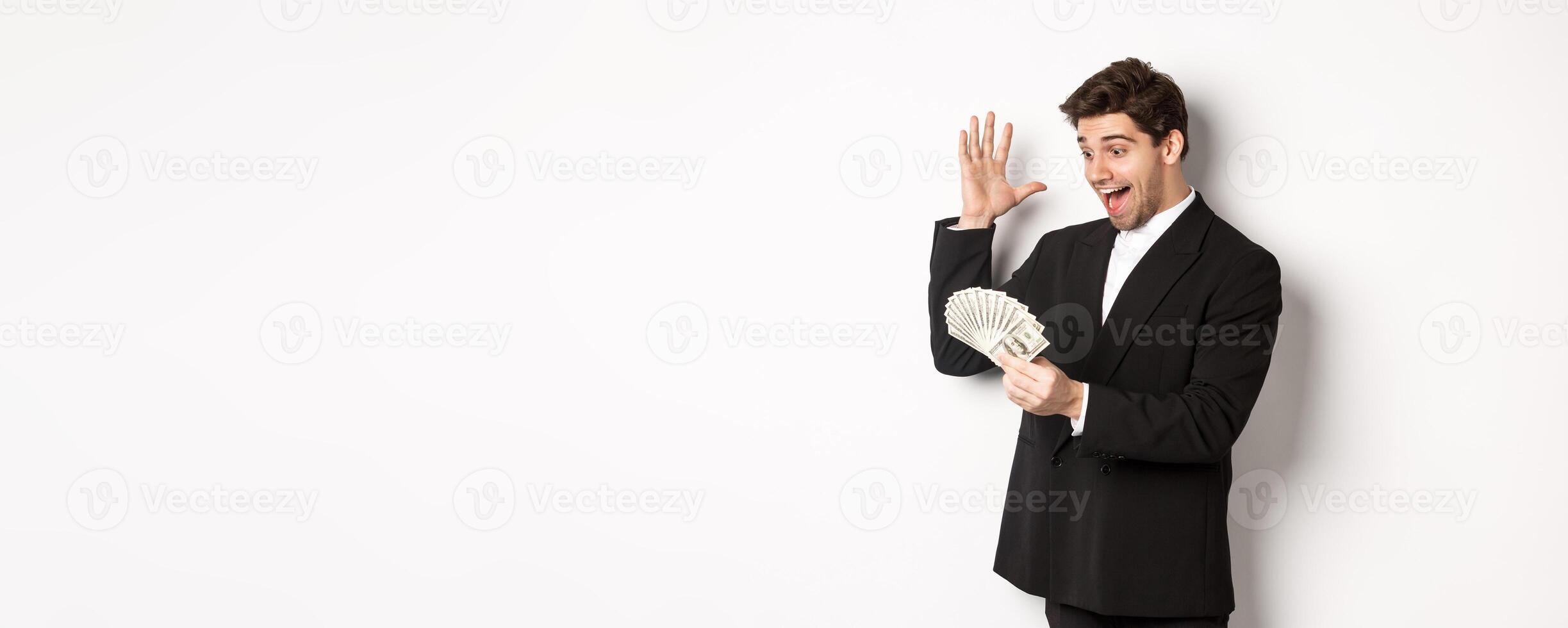 Image of successful businessman in black suit, looking at money and triumphing, earn cash, standing over white background photo
