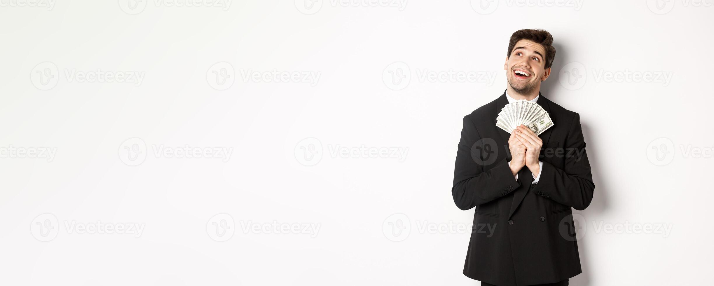 Image of handsome dreamy man in black suit, holding money and looking at upper left corner, thinking about shopping, standing over white background photo