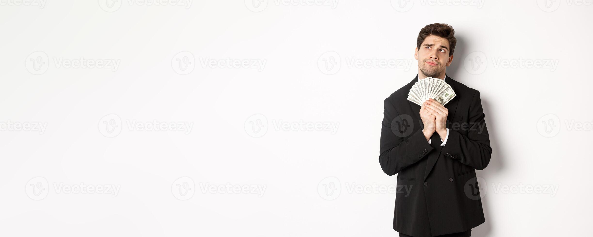 Image of handsome businessman in black suit, holding money and thinking about investment, making shopping plans, standing over white background photo
