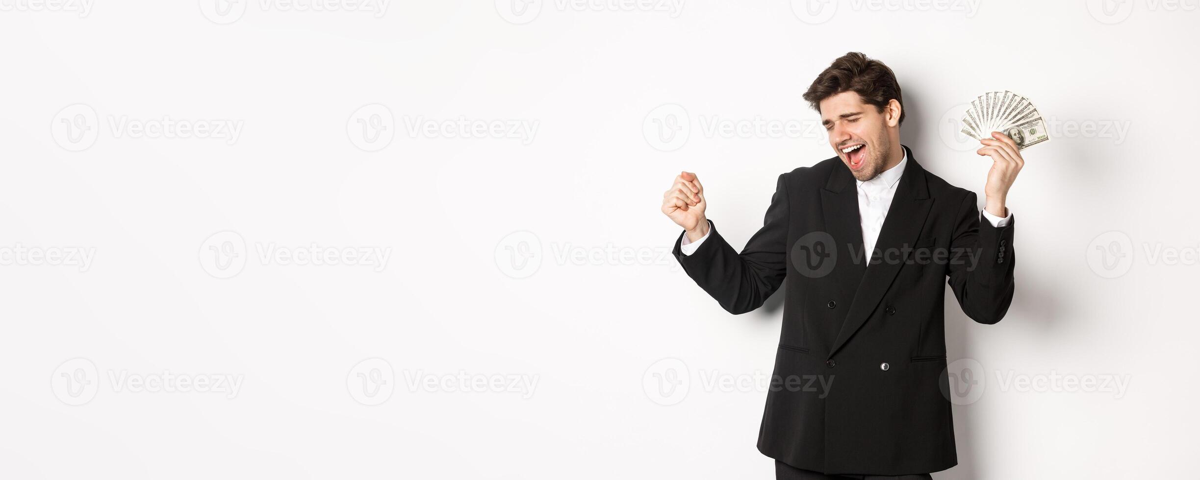 Portrait of handsome and successful businessman in suit, dancing with money, standing against white background photo