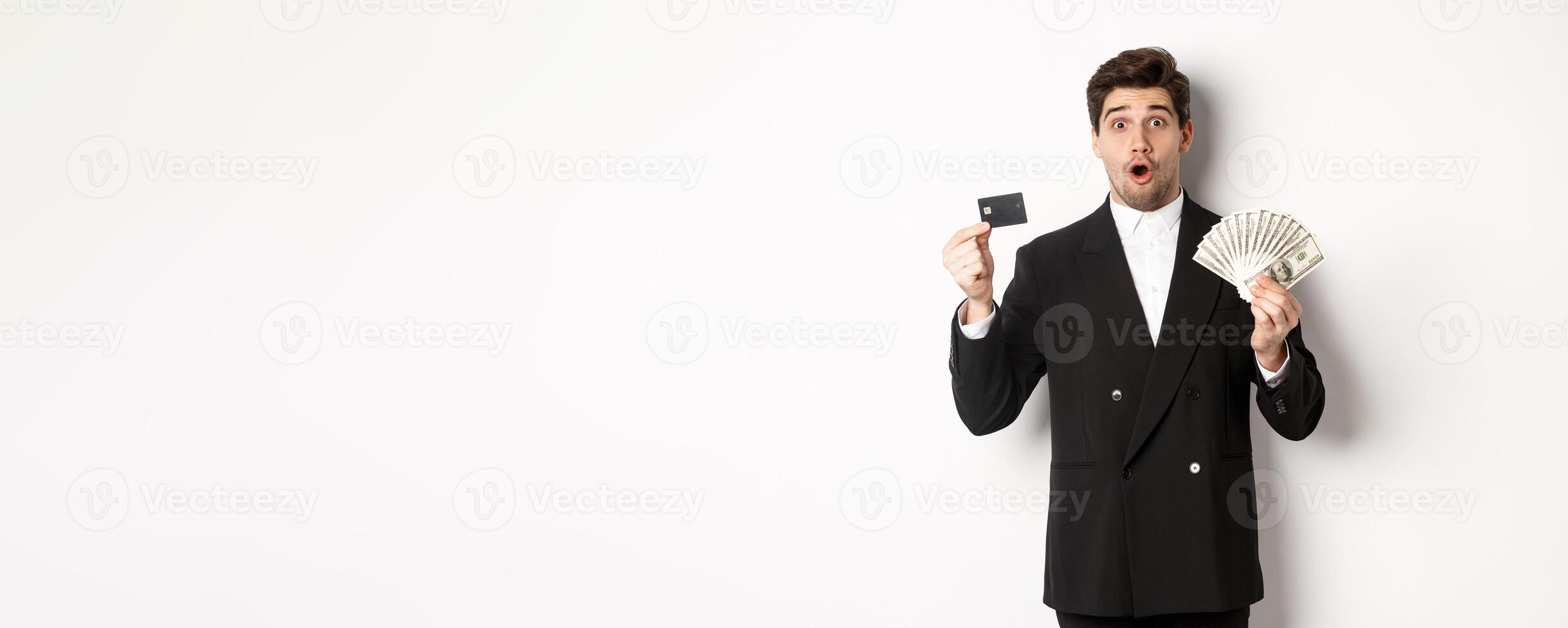 Portrait of surprised handsome man i suit, showing credit card with money, standing against white background photo