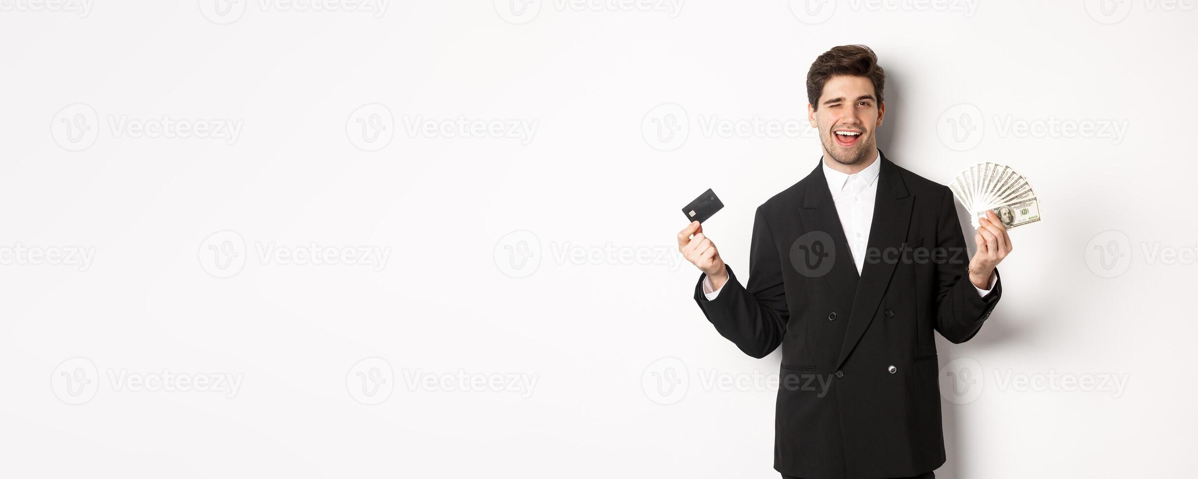 Image of confident businessman in black suit, smiling pleased and winking, holding money and credit card, standing against white background photo